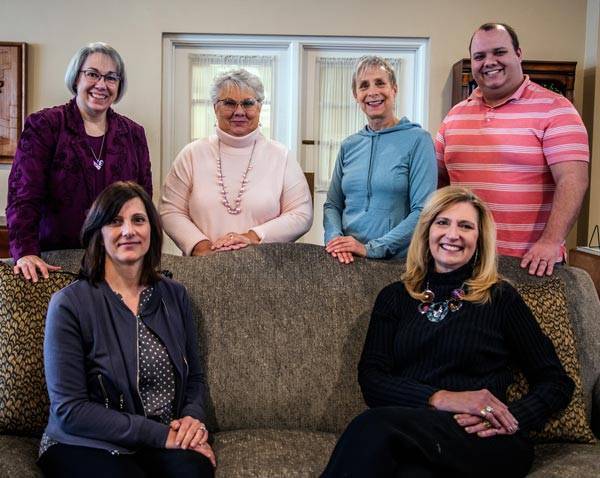 All six staff members. Four standing, and 2 sitting on a couch. Everyone is facing the camera and smiling.